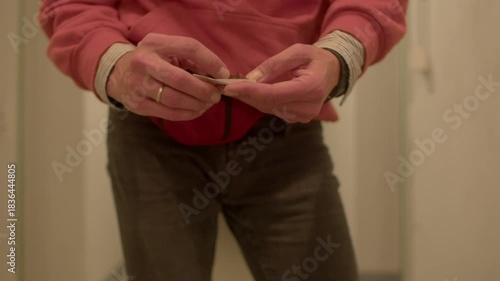 hands rolling paper closeup in narrow hallway, candid shot of fingers shaping cigarette, pink sweater and waist bag visible, focused lowlight warm tones, careful ritualistic motion and subtle tension