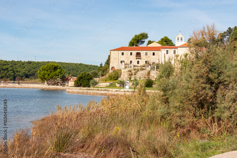 Obraz premium The historic stone Monastery of St. Euphemia stands on a hill overlooking the calm bay, framed by tall coastal reeds and lush pine forests under a blue sky on the sunny coast of Rab Island, Croatia.
