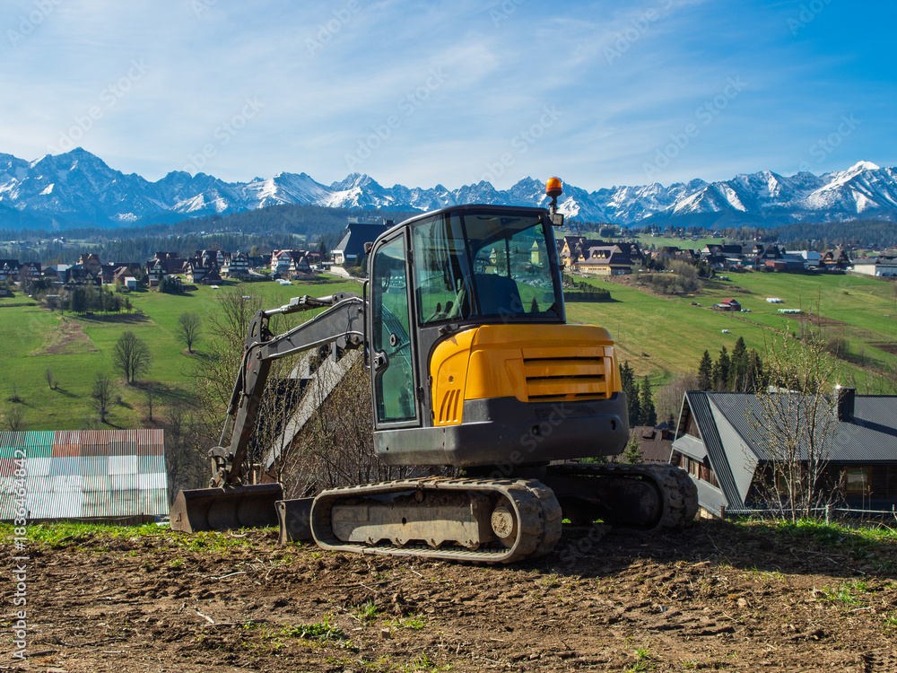 Obraz premium A small yellow tracked excavator works on a construction site, moving soil on a sunny day with the picturesque Polish Tatra Mountains and traditional rural houses in the background.