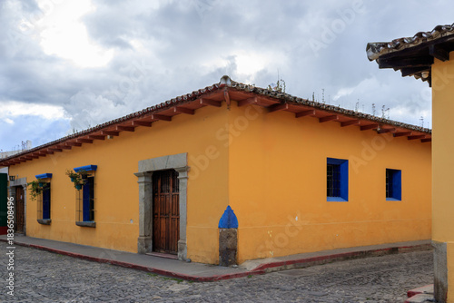 Bright yellow building with blue accents captures the charm of a colonial street in Antigua, Guatemala under a cloudy sky