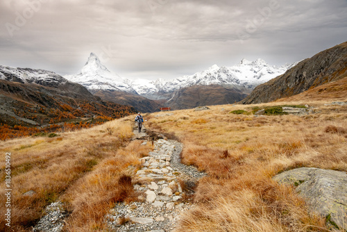 Hiker Climbs Hill Through Dry Grassland While The Matterhorn Rises To The Cloudy Sky
