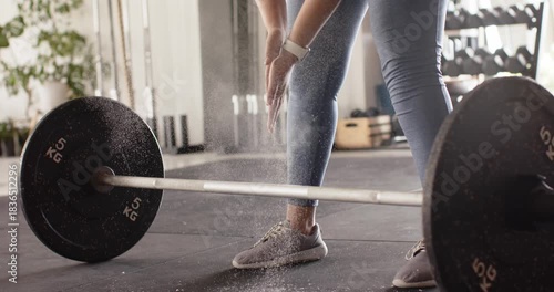 Hands clapping chalk over barbell on gym floor, chalk dust drifting while legs bracing for deadlift