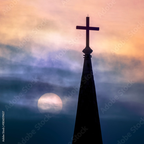 silhouette of cross on steeple top in front of dramatic sky with sun shining behind dark clouds