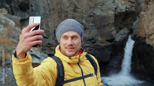 Happy male traveler taking a selfie with his smartphone while enjoying the stunning view of a waterfall, then sharing the picture on social media during his adventure in the mountains