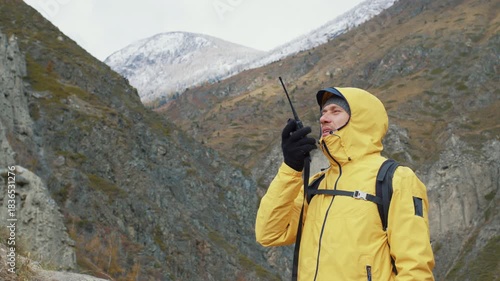 Determined male tourist in a yellow hooded jacket with a backpack talking on a portable radio while exploring a remote mountain range with snowy peaks and a cloudy sky in the background