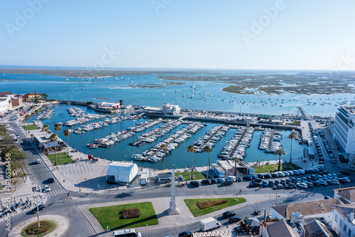 People visit the marina in Faro, Portugal with many boats docked and Ria Formosa in the distance. The sun shines brightly over the old town area