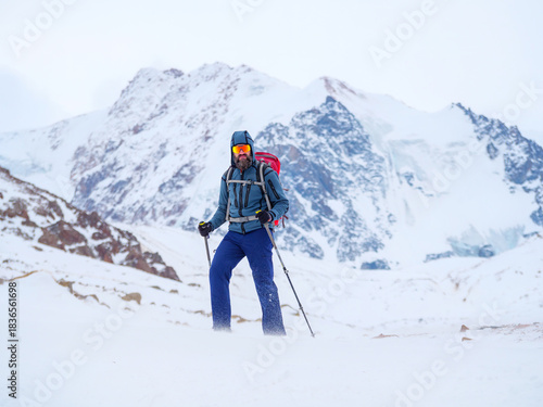 Bearded man mountain tourist with trekking poles is on a snow-covered alpine ridge in bad weather during a winter expedition