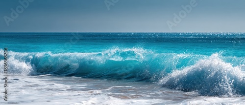 Majestic turquoise and blue ocean wave breaking with white foam and spray on a sandy beach under a clear sky, capturing the raw power and beauty of the sea.