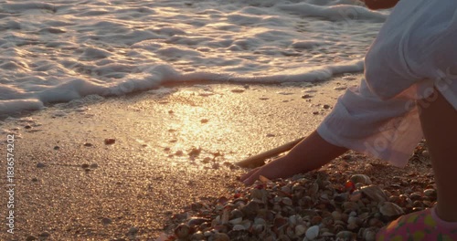 A child squats on a sandy beach, collecting shells as gentle ocean waves lap the beach during a beautiful golden hour sunset, reflecting the sunlight on the water. The child plays in the surf.