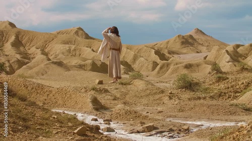 Wide Shot of a human representation of Christ in traditional biblical-style clothing, walking through an arid desert landscape, surrounded by sculpted sand formations and warm golden light. Cinematic