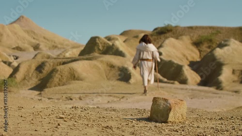 Long Shot of a Solitary Traveler in simple traditional attire crosses an arid, sunlit terrain, using a wooden staff while walking through a quiet and rugged wilderness. Christian and spiritual concept