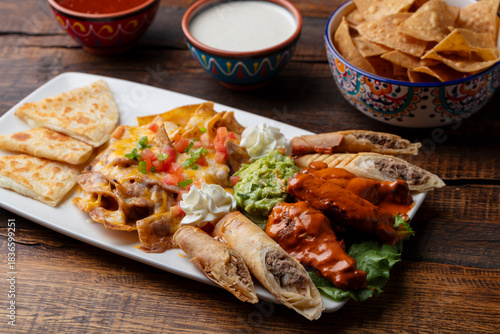 Mexican food appetizer plate with chips, salsa, nachos, taquitos, quesadillas, guacamole, chicken wings with sauce, and sour cream on a white plate. 