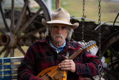 Man With Mustache Wearing Cowboy Hat and Red Flannel Shirt Holding Mandolin String Instrument Sitting On Rustic Swing In Front of Old Wooden Wagon Wheel Outside of  Rural Saloon Bar
