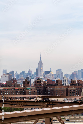 Empire State Building and midtown Manhattan skyline seen from the Brooklyn Bridge, featuring urban architecture and the iconic New York City cityscape under a bright sky