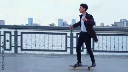 Man in a business suit is rolling on skateboard along embankment, pushing off with his foot, enjoying speed. Guy practicing skateboarding on esplanade. Active, sportive lifestyle, hobby