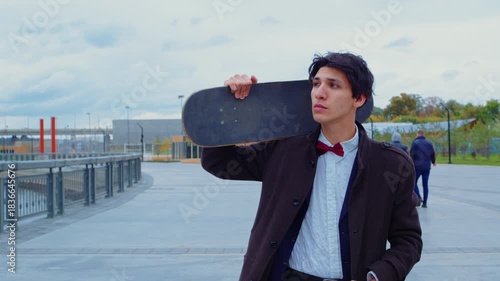 Handsome guy portrait on urban street. Boy in coat with skateboard walks on embankment, looks at camera. Office worker doing skateboarding on esplanade after work. Skateboarder, extreme sport