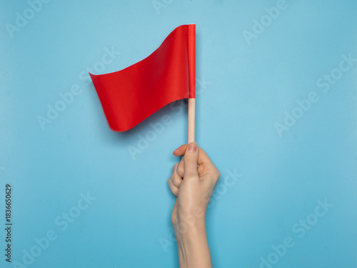Hand holding a small red flag against a blue background. Symbol of alert, warning, danger sign or protest. Minimalist conceptual image.