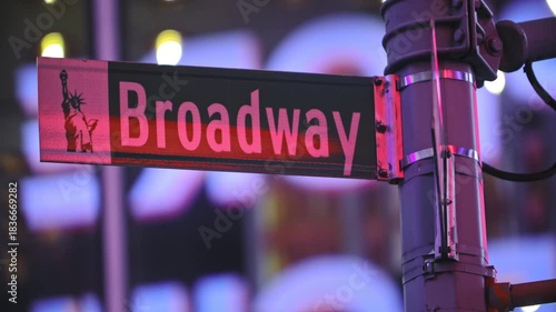 A vibrant, close-up shot of the iconic Broadway street sign in Manhattan, New York City, illuminated against a blurred background of city lights and billboards at night. 