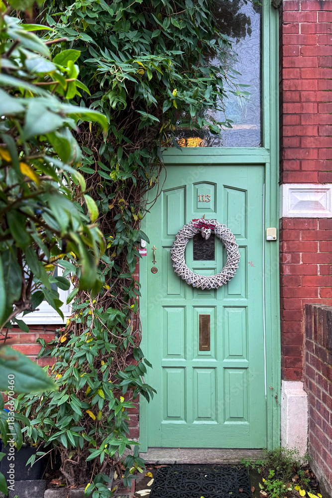 Fototapeta premium London, UK, 6 December 2025, A classic, bright green painted wooden front door of a brick house with a wicker Christmas wreath, partially covered by climbing green vines and foliage.