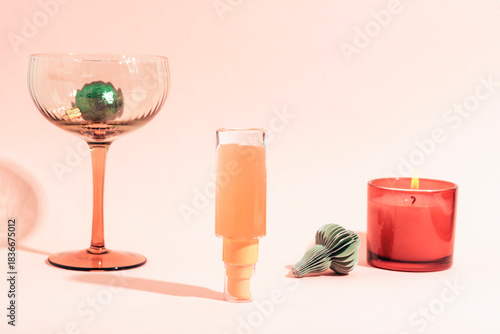 Cosmetic serum bottle standing upside down, candle, wineglass and Christmas ornaments on pink background. Holiday-themed beauty products, mockup