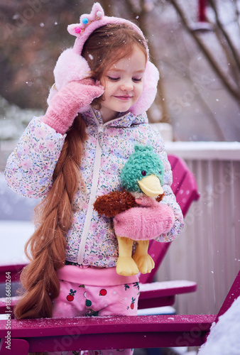 Winter Portrait of Smiling Little Girl in Fluffy Earmuffs Holding Plush Duck Toy During Gentle Snowfall – Cozy Pastel Childhood Moment While Playing Outdoor
