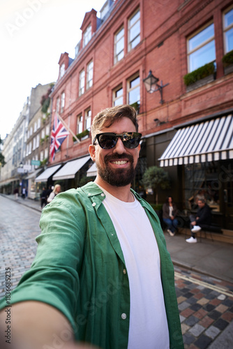 Vertical. Smiling man taking a selfie looking at camera on a London street with a Union Jack and striped awnings behind him. Casual travel moment, daylight, urban vibes and cheerful tourism energy.