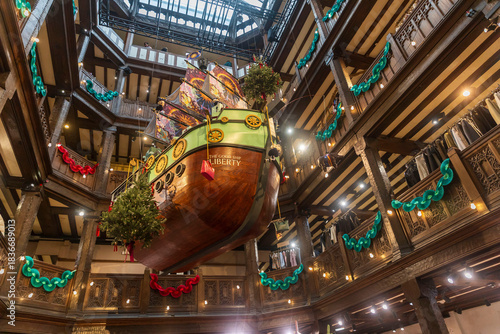 London, UK, An upward view of the “Good Ship Liberty” Christmas installation, a large wooden ship suspended in Liberty London’s timber-framed atrium, adorned with festive garlands and lights.