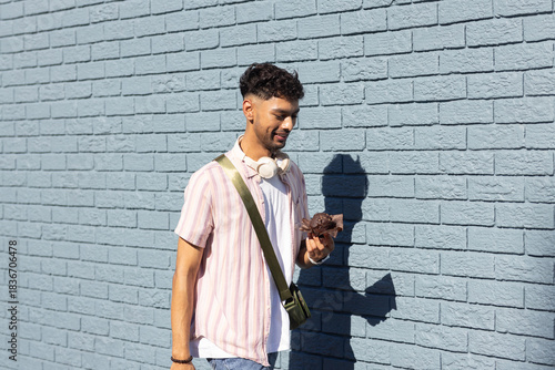 Indian man walking alongside blue wall on sidewalk holding dried leaves and wearing headphones