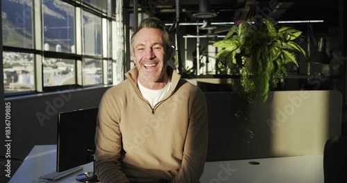 Mature adult male gazing out window, turning head, smiling and laughing at white desk with computer