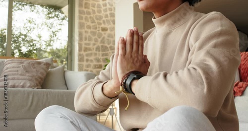 Adult Asian man bringing palms into prayer then lowering hands into mudra meditating beside sofa