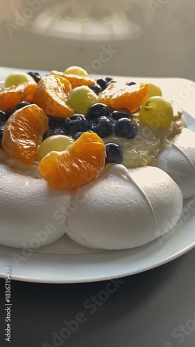 Close-up of a Pavlova meringue dessert topped with creamy filling and fresh berries, including red currants, blueberries and citrus slices. Bright directional light enhances textures and colors.
