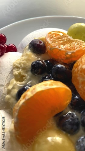 Close-up of a Pavlova meringue dessert topped with creamy filling and fresh berries, including red currants, blueberries and citrus slices. Bright directional light enhances textures and colors.