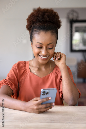 Happy young Black mobile phone user woman browsing Internet, social media on mobile phone, reading message, smiling, sitting at table, using smart app, web service. Vertical shot