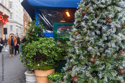 Fototapeta Naklejka Na Ścianę i Meble -  London, UK, 6 December 2025, A close-up of a snow-flocked Christmas tree with pine cones next to a small blue-awning cafe on a London street, with blurred pedestrians in the background.