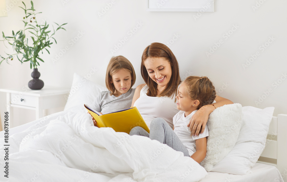 Obraz premium Cheerful mother and children spend leisure time together in bed, reading a book at home. They share family bonding in the bedroom, enjoying quiet moments of togetherness and relaxation.