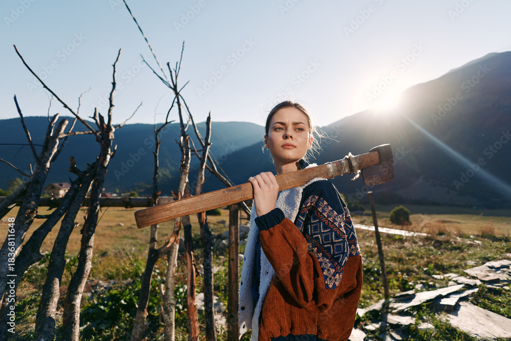 Obraz premium Woman carrying an axe on her shoulder in countryside near a rustic fence with mountains and sunrise in background, wearing a patterned sweater and looking confident.