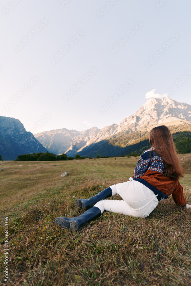 Obraz premium Woman sitting on grassy meadow facing mountains and nature landscape in autumn, wearing cozy sweater, jacket and boots, relaxing outdoors with long hair and peaceful view