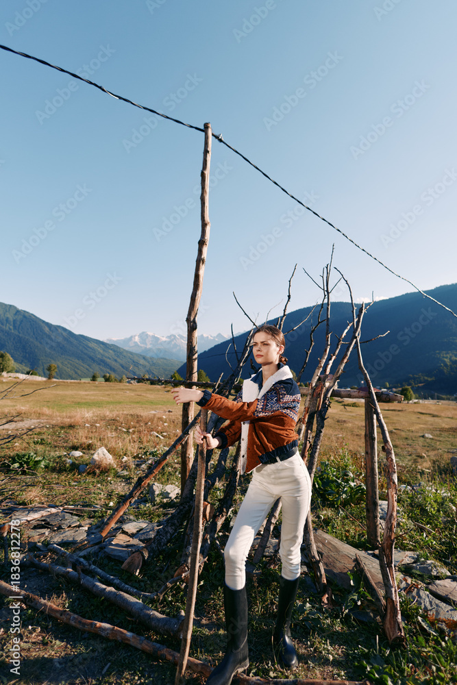 Obraz premium woman fence mountains field boots jacket rural outdoors portrait of young model leaning on handmade wooden fence in alpine meadow, casual riding style, natural light and distant peaks