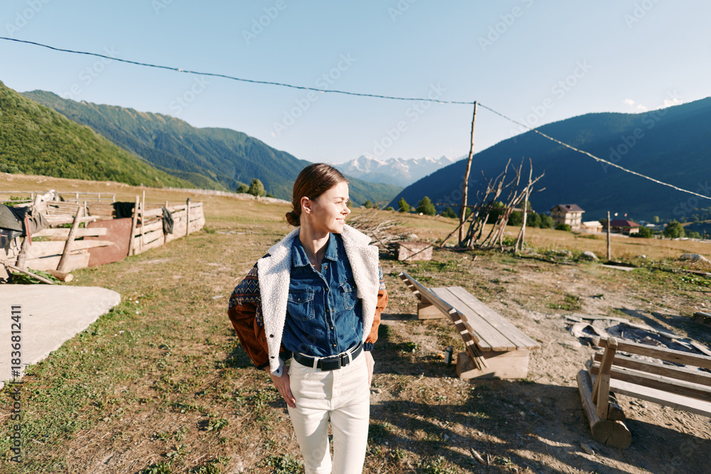 Obraz premium Woman in countryside with mountains and meadow, smiling outdoors while wearing a backpack and casual clothes. Rural landscape in sunlight, relaxed portrait and travel lifestyle scene.