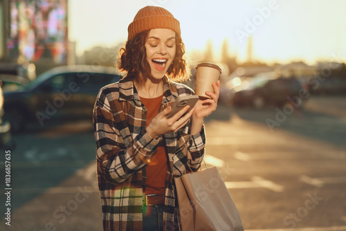Fotografija Woman with coffee and phone, smiling while shopping in urban sunlight at golden hour glow