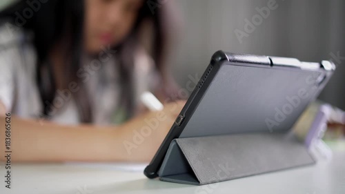 closeup tablet with blur asian child student study online or young kid girl writing or notes on paper with computer and doing homework or people learn from homeschool and back to school by smartphone