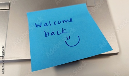 Hand written welcome back message with smiley face from friend on blue sticky note left on laptop in office environment 