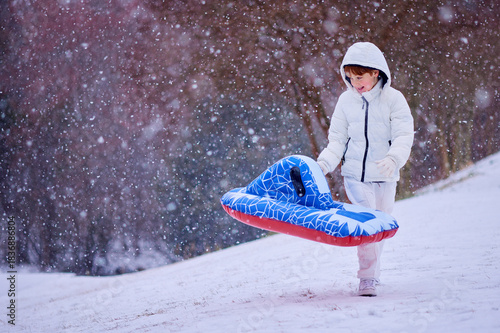 Happy Boy Walking Up Snowy Hill with Inflatable Sled - Winter Adventure in Falling Snow. Fun Seasonal Ride