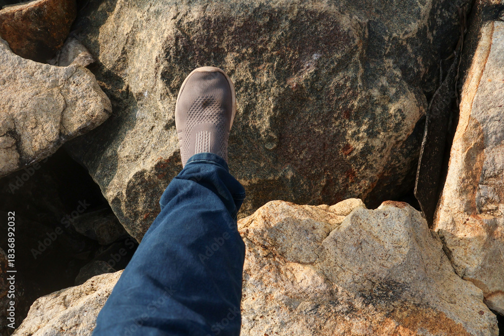 Fototapeta premium Person walks carefully on large coastal boulders of the Shangumugham Beach sea wall, wearing casual shoes and jeans in warm evening light.
