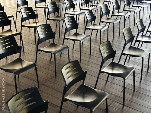 Empty black chairs arranged on a wooden floor in a spacious, well-lit room, creating a pattern of lines and shadows, evoking a sense of readiness for an event or meeting