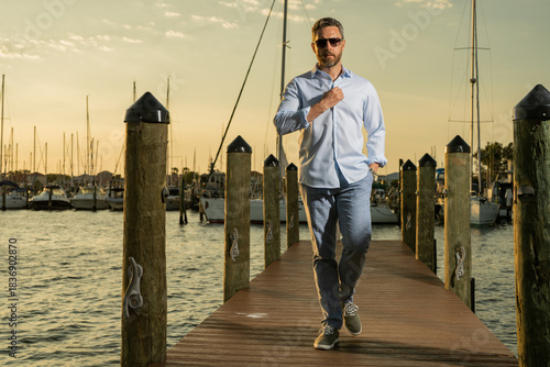 Rich fashion business man dreaming and thinking of yacht. Well-dressed fashion man in fashionable eyeglasses and shirt posing near yacht. Male model in yacht club. Portrait of stylish man. Male