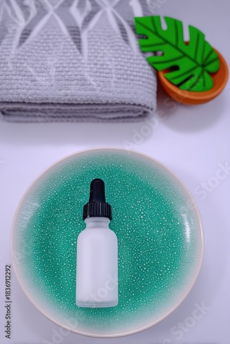 A White Frosted Dropper Bottle of Face Oil Sits on a Textured Green Ceramic Plate Next to a Folded Gray Bath Towel and a Green Leaf Decoration on a White Surface