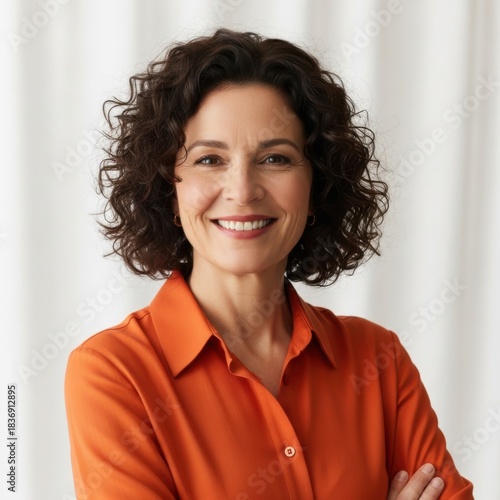 Friendly smiling woman portrait wearing orange shirt with arms crossed and curly dark hair in studio lighting with blurred background isolated on white background