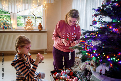 Little school girl and mother decorate traditional Christmas tree with vintage decorations and toys at home