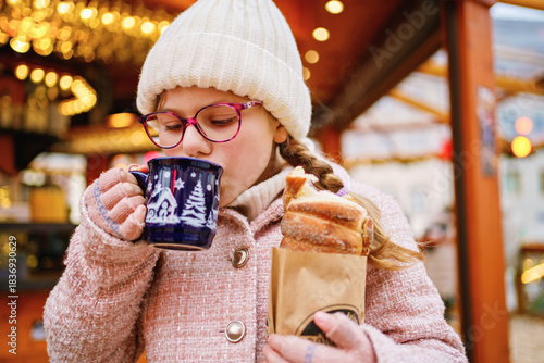 Little cute school girl eating sweet bread called Baumstriezel and drink hot chocolate or punch on German Christmas market. Happy child on traditional family market in Germany,